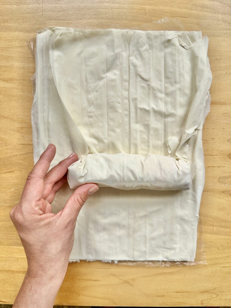 Phyllo dough rolled over the chicken and a hand is holding the roll in place over a wooden table